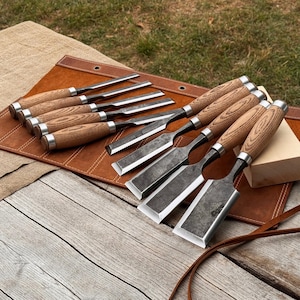 Peut inclure: Un ensemble de ciseaux à bois avec des manches en bois brun clair et des lames en métal sombre texturé, disposés dans une trousse à outils en cuir brun. Les ciseaux varient en largeur et possèdent des viroles en métal poli.