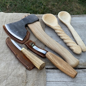 Puede incluir: Una colección de herramientas de carpintería hechas a mano, que incluye un hacha pequeña con funda de cuero, un cuchillo con funda de cuero, un cuchillo curvo para tallar y dos cucharas de madera. Las herramientas son de madera y metal.