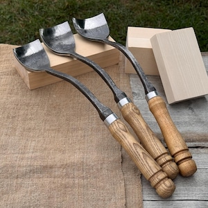 May include: Four wood carving gouges with curved, silver-colored blades and wooden handles. The tools are arranged on a tan cloth surface, with two small wooden blocks in the background. The handles have a light brown finish.