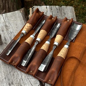 May include: A set of five wood carving gouges with polished metal blades and light-colored wooden handles, each secured in an individual brown leather sleeve within a roll-up leather tool organizer featuring white stitching and a tie closure.