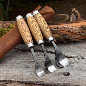 May include: Three hand-forged metal carving tools with light-colored, wood-grained cylindrical handles and silver-toned metal ferrules. Each tool features a curved, sharpened blade with a distinct stamped logo near the base of the metal shank.