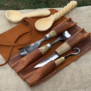 May include: A collection of wood carving tools and wooden spoons. The tools include a knife, gouges, and a hook knife, all with wooden handles and leather sheaths. Two wooden spoons are also visible, one with a twisted handle.