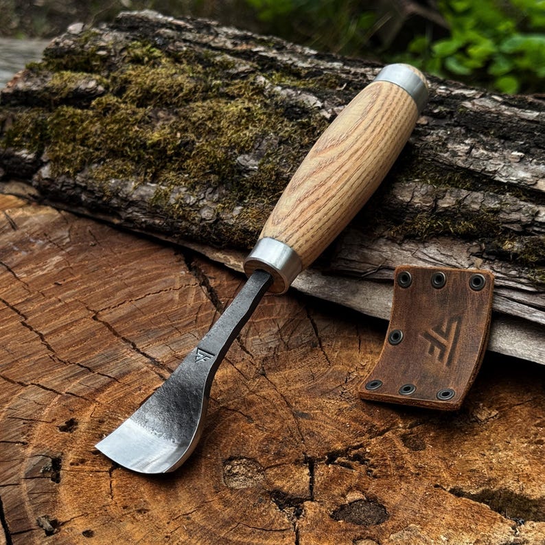 May include: A wood carving tool with a curved metal blade and a light brown wooden handle featuring silver metal ferrules. Beside the tool lies a small, curved brown leather sheath with black rivets and a stamped logo featuring the letters TF.