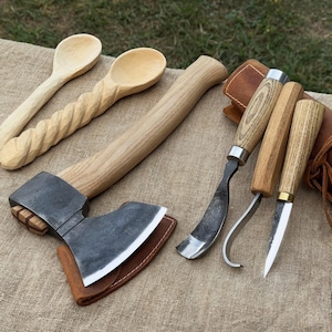 Puede incluir: Una colección de herramientas para trabajar la madera, que incluye dos cucharas de madera, un hacha pequeña con funda de cuero y tres cuchillos para tallar. Las herramientas son de madera y metal, con un acabado natural.