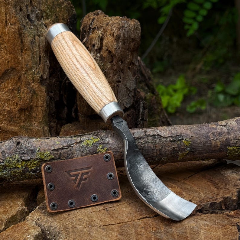 Pu&ograve; includere: Un coltello a uncino con manico in legno chiaro e lama in acciaio curva. Accanto all'attrezzo si trova un fodero rettangolare in pelle marrone con un logo stampato con le lettere TF e sei occhielli metallici lungo i bordi della pelle.