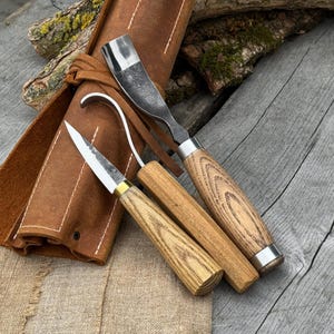 May include: A set of three wood carving tools with wooden handles and metal blades, alongside a brown leather tool roll. The tools include a curved gouge, a hook knife, and a straight chisel. The handles have a natural wood grain finish.