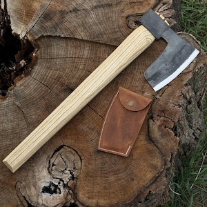 May include: A hand axe with a long, light-colored wooden handle and a dark, forged metal head featuring a sharpened silver edge. Beside the axe head lies a small, brown leather sheath with a single metal snap button closure for protection.