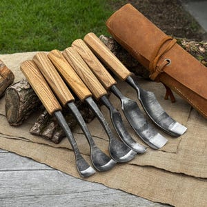 May include: A set of six wood carving tools with light brown wooden handles and dark, curved metal blades. The tools are arranged in a row, accompanied by a brown leather roll-up case with a tie closure, resting on a textured fabric surface.