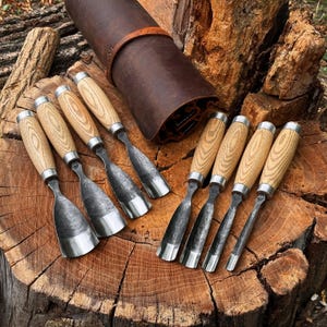 May include: A set of eight wood carving chisels with light-coloured wooden handles and dark metal blades, arranged alongside a brown leather tool roll. The chisels feature varying blade widths and shapes, including flat, curved, and gouge styles.