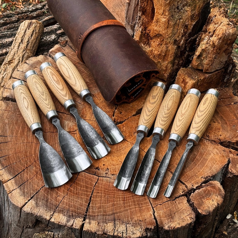 May include: A set of eight wood carving chisels with light-coloured wooden handles and dark metal blades, arranged alongside a brown leather tool roll. The chisels feature varying blade widths and shapes, including flat, curved, and gouge styles.