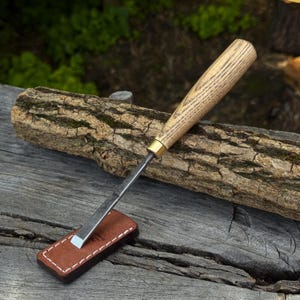 Puede incluir: Un formón para madera con hoja de metal pulido, virola de latón y mango de madera texturizada. La punta afilada del formón descansa sobre una pequeña funda rectangular de cuero marrón con costuras blancas y un logotipo grabado en relieve.