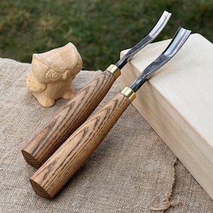 May include: Two wood carving gouges with curved blades and wooden handles. The handles have a natural wood grain pattern and are accented with brass ferrules. A small, carved wooden owl figurine is also visible.