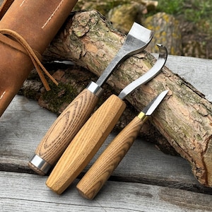 May include: Three wood carving tools with wooden handles and metal blades are displayed on a wooden surface. The tools include a straight chisel, a curved gouge, and a small knife. A leather tool roll is visible in the background.