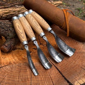 Pu&ograve; includere: Quattro sgorbie per intagliare il legno con manici in legno chiaro e ghiere in metallo argentato. Le lame in acciaio sono curve e di colore scuro. Un rotolo portautensili in pelle marrone con logo stampato e laccio &egrave; posizionato dietro.