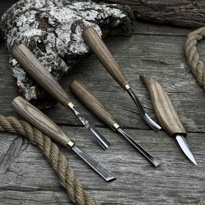 May include: A collection of wood carving tools with wooden handles and metal blades. The tools are arranged on a weathered wooden surface, with a rope and a piece of bark in the background. The handles have a natural wood grain finish.