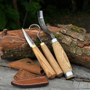 May include: Three woodworking tools with light-coloured wooden handles and metal blades, including a straight knife, a hook knife, and a gouge. Each tool features a small logo on the blade. Three brown leather sheaths are included in the set.
