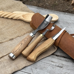 May include: A set of wood carving tools with wooden handles and metal blades, including a gouge, a hook knife, and a detail knife. The tools are displayed with a leather tool roll and a carved wooden spoon. The wood has a natural grain.