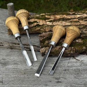 May include: A set of four wood carving chisels featuring light-colored wooden mushroom-shaped handles with silver metal ferrules and polished steel blades of varying shapes, including gouge and flat profiles for woodworking tasks.