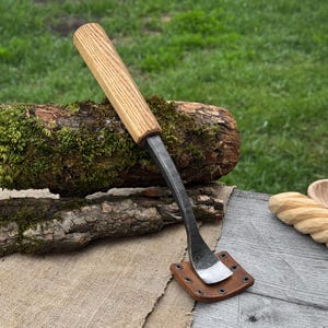 Op de afbeelding: Een handgesmede trekmes met een houten handvat en een gebogen stalen lemmet. Het lemmet wordt beschermd door een bruine leren schede. Het gereedschap rust op een stuk hout met mos.