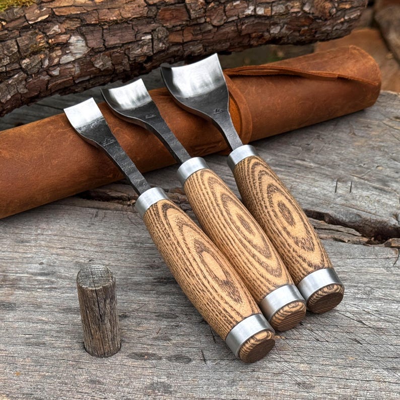 May include: Three metal wood carving gouges with curved steel blades and light brown wooden handles featuring a distinct grain pattern. Each tool has a silver metal ferrule at the top and bottom of the handle, and they are arranged in a row.