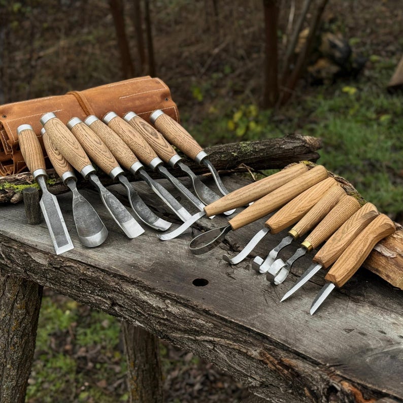 Pode incluir: Um conjunto de ferramentas de entalhe em madeira com cabos de madeira e l&acirc;minas de metal, dispostas sobre uma superf&iacute;cie de madeira envelhecida. Um rolo de ferramentas de couro castanho &eacute; vis&iacute;vel no fundo, sugerindo um conjunto para trabalhar madeira ou esculpir.