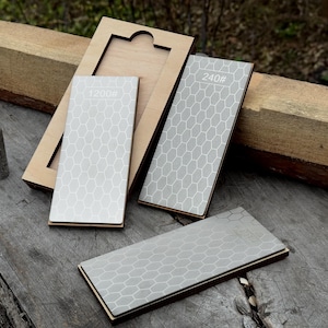 May include: A set of sharpening stones with wooden bases. The stones are gray with a white honeycomb pattern and the numbers 240#, 400#, and 1200# printed on them. One stone is inside a wooden box.