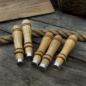 Könnte beinhalten: Sechs hölzerne Flaschenverschlüsse. Jeder Verschluss hat einen hellbraunen, zylindrischen Griff und einen silberfarbenen Metallsockel. Sie sind auf einer verwitterten Holzoberfläche mit einem Seil im Hintergrund angeordnet.