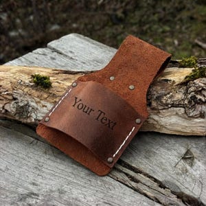 May include: A brown leather tool holder with the text "Your Text" engraved on the front. The holder is secured with rivets and white stitching. The background is a weathered wooden surface.
