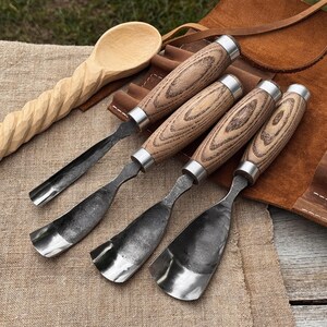 Könnte beinhalten: Eine Sammlung von Holzschnitzwerkzeugen, darunter vier Hohleisen mit Holzgriffen und Metallklingen sowie zwei Holzlöffel. Die Hohleisen haben ein silbernes Metallband am Griffansatz. Die Werkzeuge liegen auf einer beigen Oberfläche.