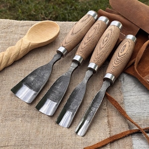 May include: A set of four wood carving gouges with wooden handles and metal blades. The gouges are arranged on a tan fabric surface, with a carved wooden spoon in the background. A leather tool roll is visible on the right.