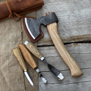 May include: A collection of woodworking tools, including a small axe with a wooden handle, a leather sheath, and three carving knives. The axe head is dark metal, and the handles of the tools are light-colored wood. The tools are arranged on a wooden surface.