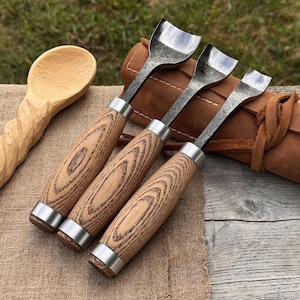 May include: Three wood carving gouges with wooden handles and metal blades are displayed. The handles have a light brown wood grain pattern, and the blades are silver. A carved wooden spoon is visible in the background, along with a leather pouch.