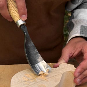 May include: A wood carving gouge with a curved, dark metal blade and a light-colored wooden handle. The tool features a small stamped logo on the metal neck and is shown removing a thin shaving of light-colored wood from a larger wooden piece.