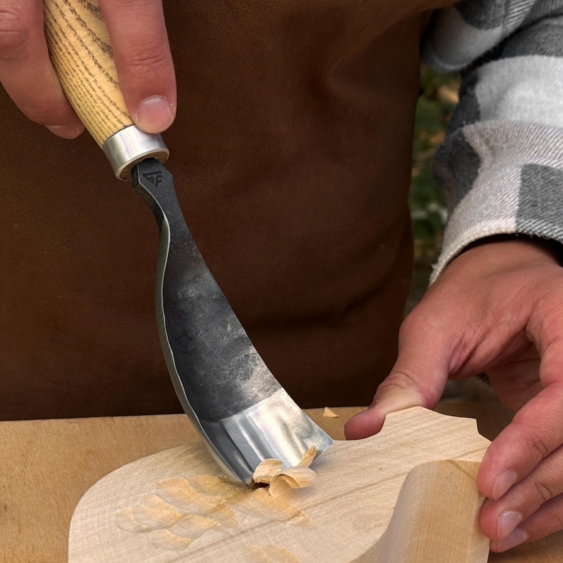 May include: A wood carving gouge with a curved, dark metal blade and a light-colored wooden handle. The tool features a small stamped logo on the metal neck and is shown removing a thin shaving of light-colored wood from a larger wooden piece.