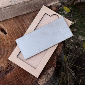 May include: A sharpening stone with a honeycomb pattern and the text "1200#" rests inside a wooden frame. The frame is light-colored, and the stone is a light gray. The items are on a wooden surface.