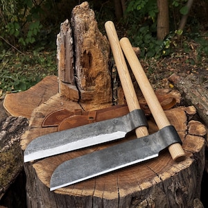 Op de afbeelding: Twee handgesmede kapmessen met donkere, getextureerde stalen bladen met een gebogen snede en rechte houten handvatten. De gereedschappen liggen op een grote, verweerde boomstronk, vergezeld van een bruine lederen schede met een klein logo.