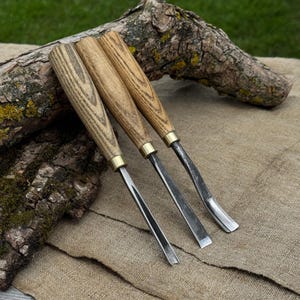 May include: Three wood carving gouges with light brown wooden handles and brass ferrules. Each tool features a distinctively curved metal blade designed for shaping wood, arranged side by side on a textured tan fabric surface.