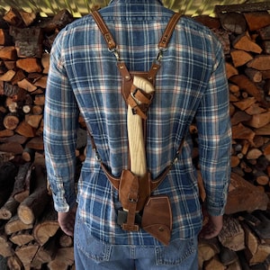 May include: A brown leather axe carrying harness with adjustable straps. The harness is worn by a person with a blue and red plaid shirt. The axe is secured in the harness with a brown leather sheath.