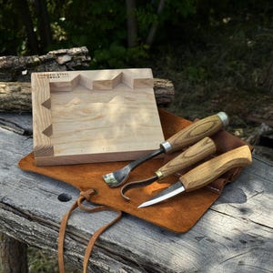 May include: A set of three wood carving tools with light brown wooden handles and metal blades, including a straight knife, a hook knife, and a gouge, resting on a brown leather roll next to a light-colored wooden block with the text FORGED STEEL TOOLS.