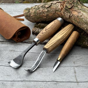 Pode incluir: Três ferramentas de entalhe em madeira com cabos de madeira e lâminas de metal são exibidas em uma superfície de madeira. Uma bolsa de ferramentas de couro marrom é visível ao fundo. As ferramentas têm diferentes formatos de lâmina.