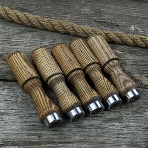 Puede incluir: Colección de cinco tapones de botella de madera clara, con bases de metal plateado. Los tapones están dispuestos sobre una superficie de madera envejecida, con una cuerda en el fondo.