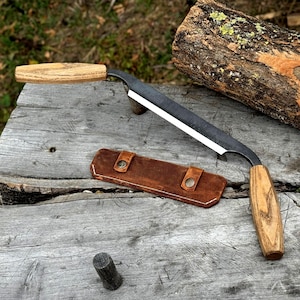 Puede incluir: Un cuchillo de tracción con una hoja oscura y mangos de madera, sobre una superficie de madera envejecida. También hay una funda de cuero marrón con dos cierres de botón. La herramienta es para trabajar la madera.