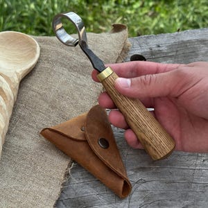 May include: A wood carving hook knife with a curved blade and wooden handle. The knife is held next to a small, brown leather pouch. The handle has a brass ferrule. The knife is designed for carving wood.