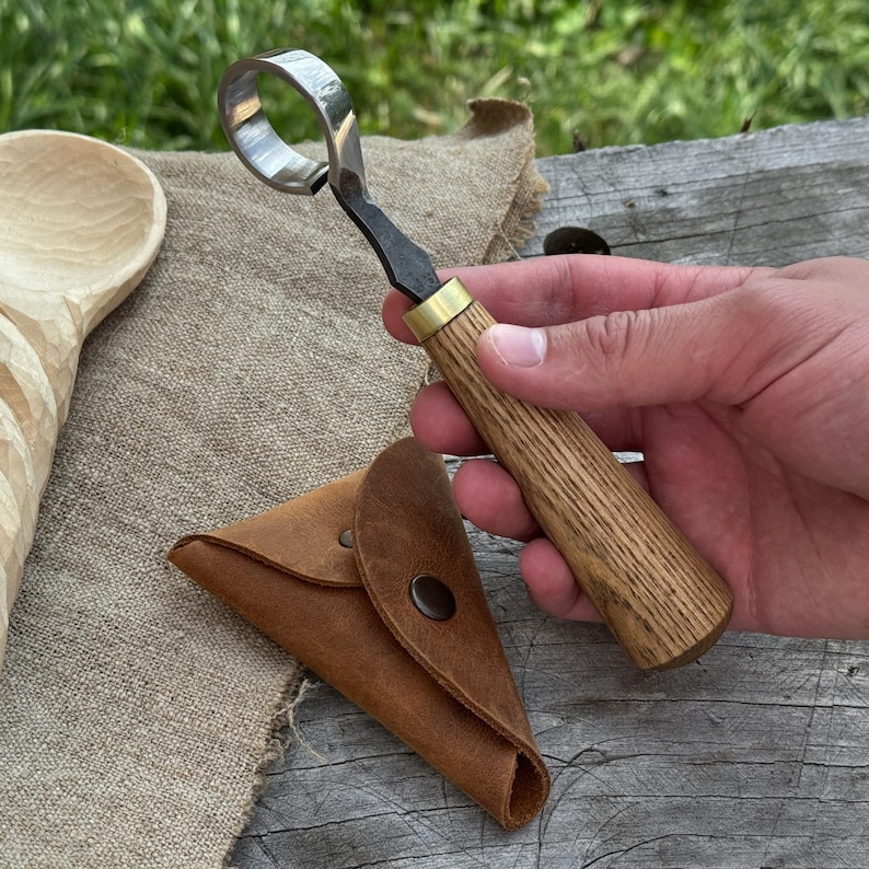 May include: A wood carving hook knife with a curved blade and wooden handle. The knife is held next to a small, brown leather pouch. The handle has a brass ferrule. The knife is designed for carving wood.