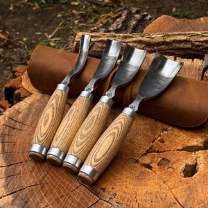 Pu&ograve; includere: Un set di quattro sgorbie per intaglio del legno con lame in metallo ricurve e manici in legno chiaro, dotate di ghiere in metallo color argento alla base di ogni manico, disposte in fila su un rotolo portautensili in pelle marrone.