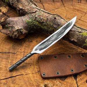 May include: A hand-forged steel knife with a pointed blade and a twisted metal handle. The blade features a dark, textured finish along the center. A brown leather sheath with metal rivets is positioned next to the knife on a wooden surface.
