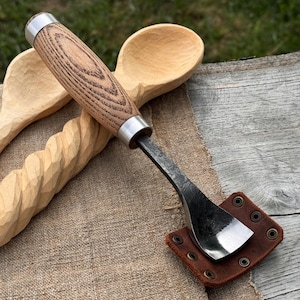 May include: A spoon carving gouge with a wooden handle and a metal blade, resting on a brown leather sheath. The handle is a light brown color with a wood grain pattern. Two wooden spoons are in the background.