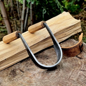 May include: A hand-forged log splitting tool with a curved, dark metal blade and two wooden handles. The tool rests on a piece of wood, with a brown leather wrist strap nearby. The background is blurred, suggesting an outdoor setting.