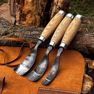 Puede incluir: Tres gubias para tallar madera con hojas de metal forjado oscuro y curvado, y mangos de madera clara con veta visible. Cada herramienta tiene una virola de metal plateado que une la hoja con el mango y un peque&ntilde;o logotipo estampado.