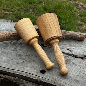 May include: Two wooden mallets with cylindrical heads and rounded handles. The mallets are made of light-colored wood, showcasing the natural grain. They are resting on a weathered wooden surface, suggesting a woodworking or crafting theme.